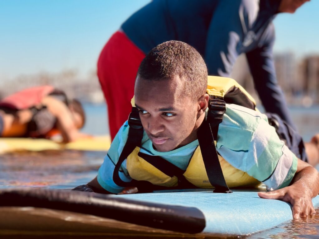A Black child lays on a paddleboard with assistance from a WATER Program lifeguard.